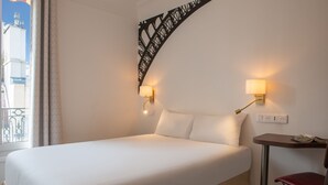 Una habitació d'hotel amb un llit gran, dues làmpades de nit, una taula petita amb una cadira i un element decoratiu a la paret.