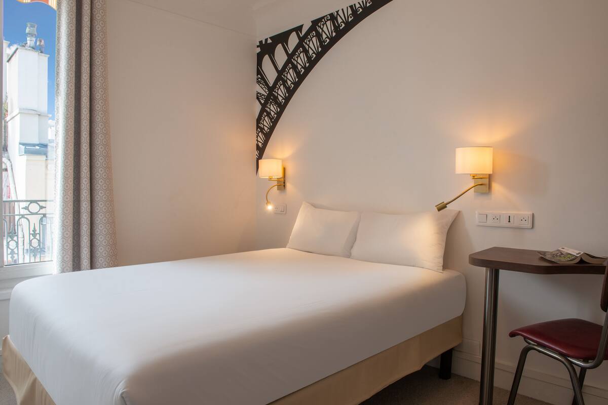 A hotel room with a large bed, two bedside lamps, a small table with a chair, and a decorative wall piece.