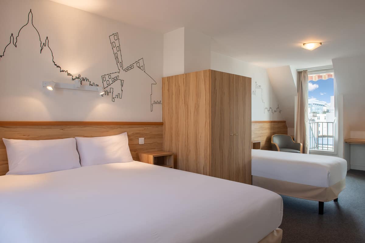 A hotel room with two beds, a wooden wardrobe, a desk, and a cityscape wall art.