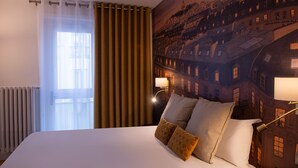 Habitació d'hotel amb un llit gran, vista a un mural urbà, una finestra amb cortines de tela fina i un radiador.