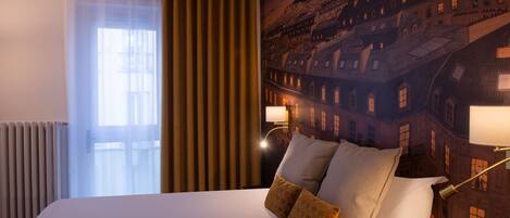 A hotel room with a large bed, a view of a cityscape mural, a window with sheer curtains, and a radiator.