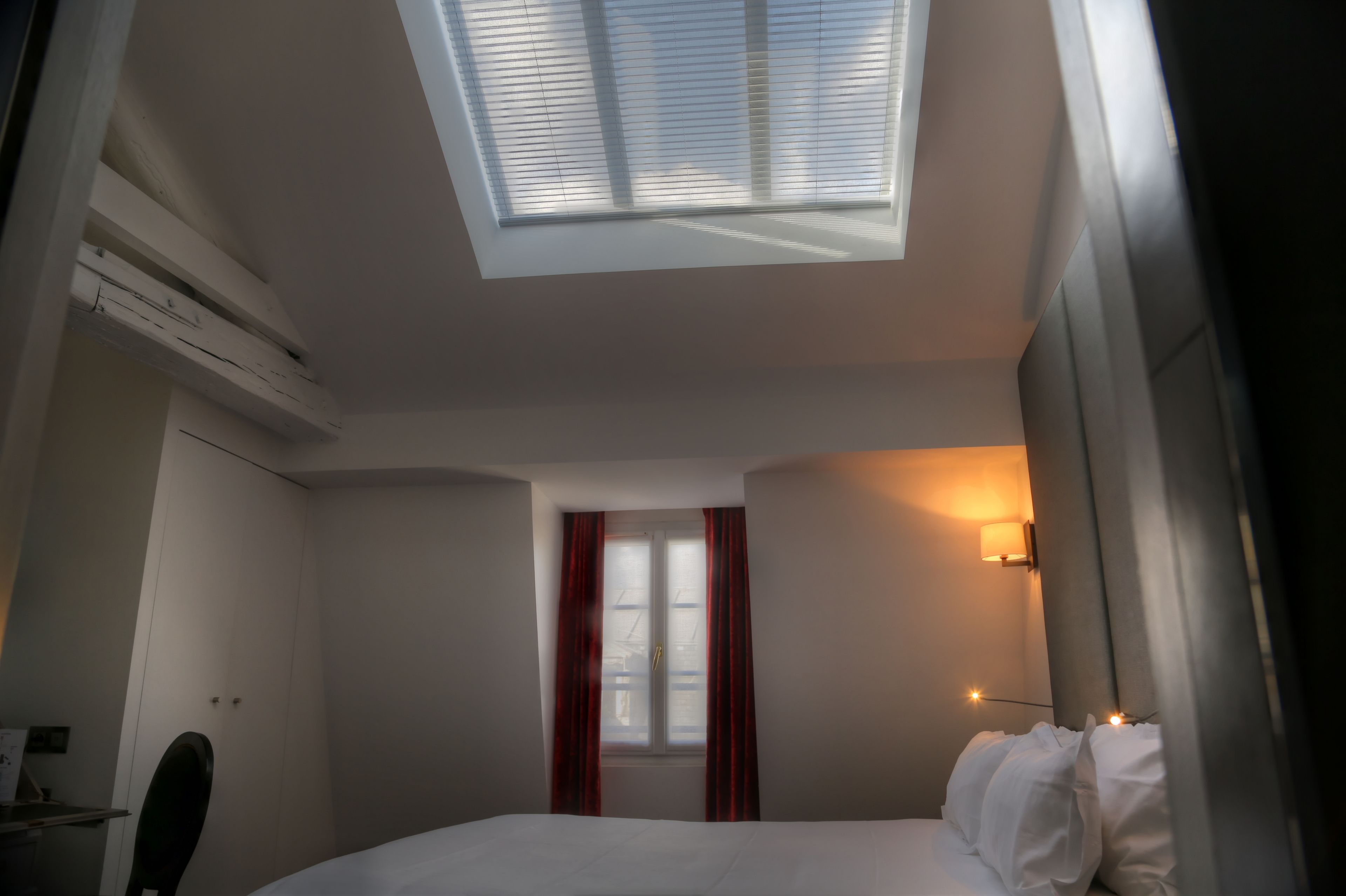 A hotel room with a skylight, a bed, a desk, and a window with curtains.