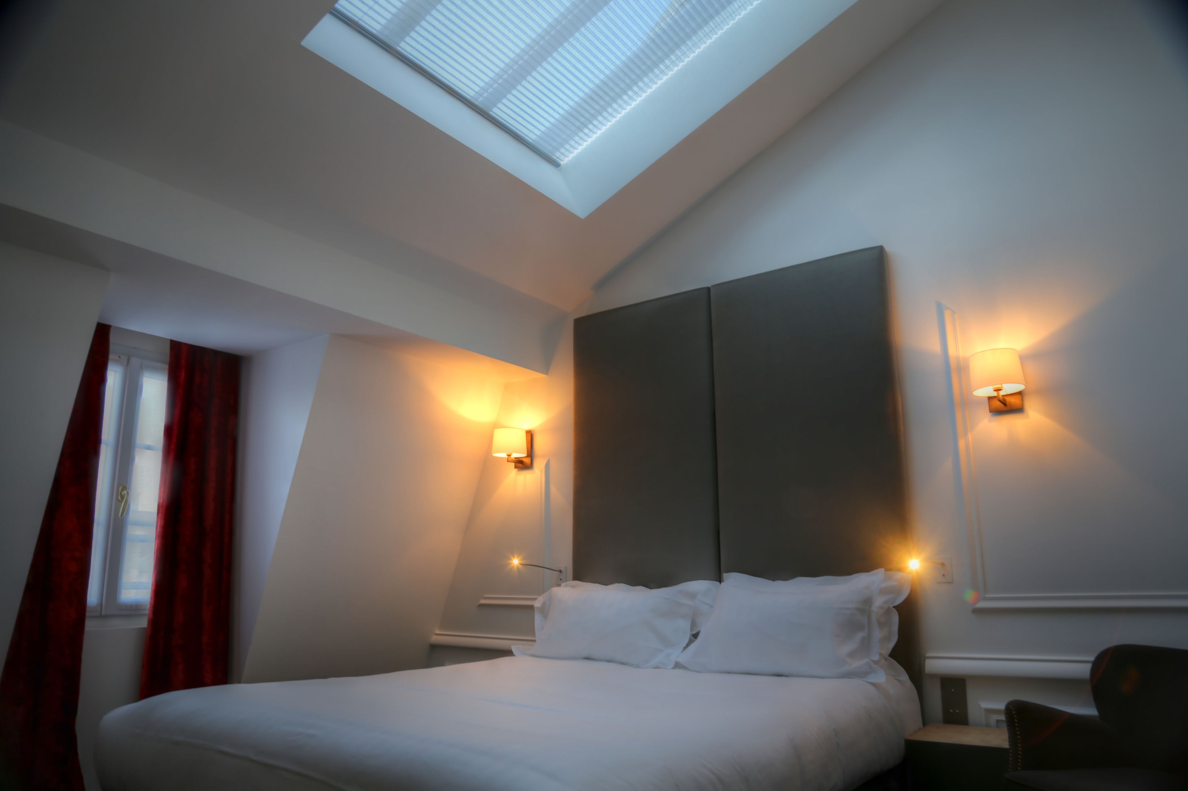 A hotel room with a skylight, a bed, two wall-mounted lamps, and a window with red curtains.