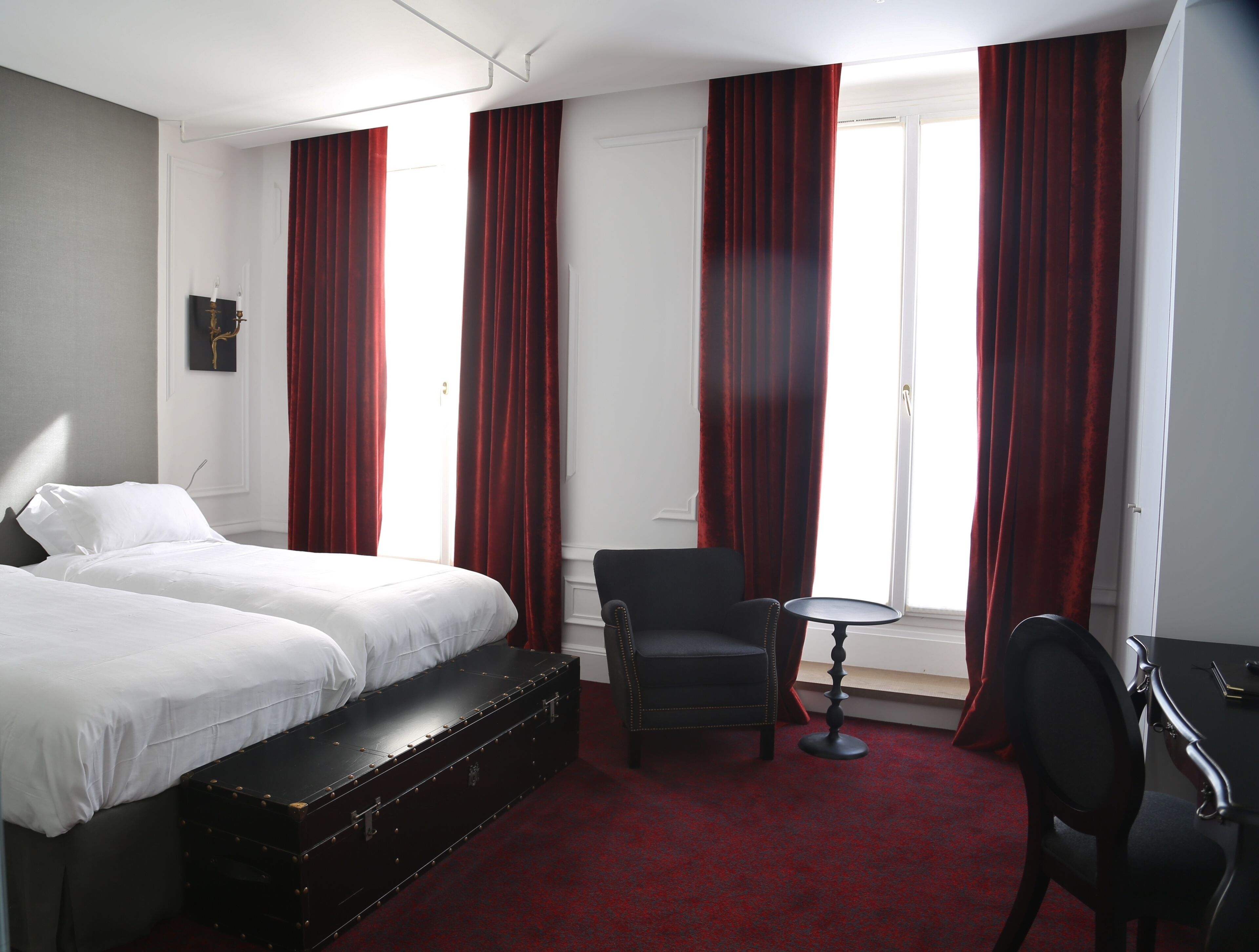 A hotel room with two beds, a desk, a chair, and a large window with red curtains.