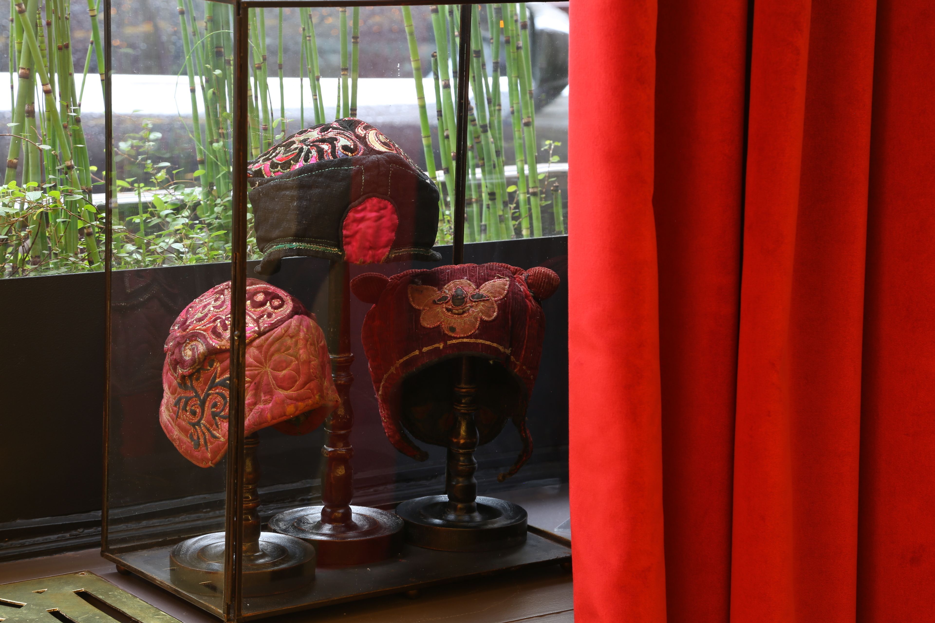 Una vitrina con tres sombreros bordados frente a una ventana con bambú visible en el exterior.