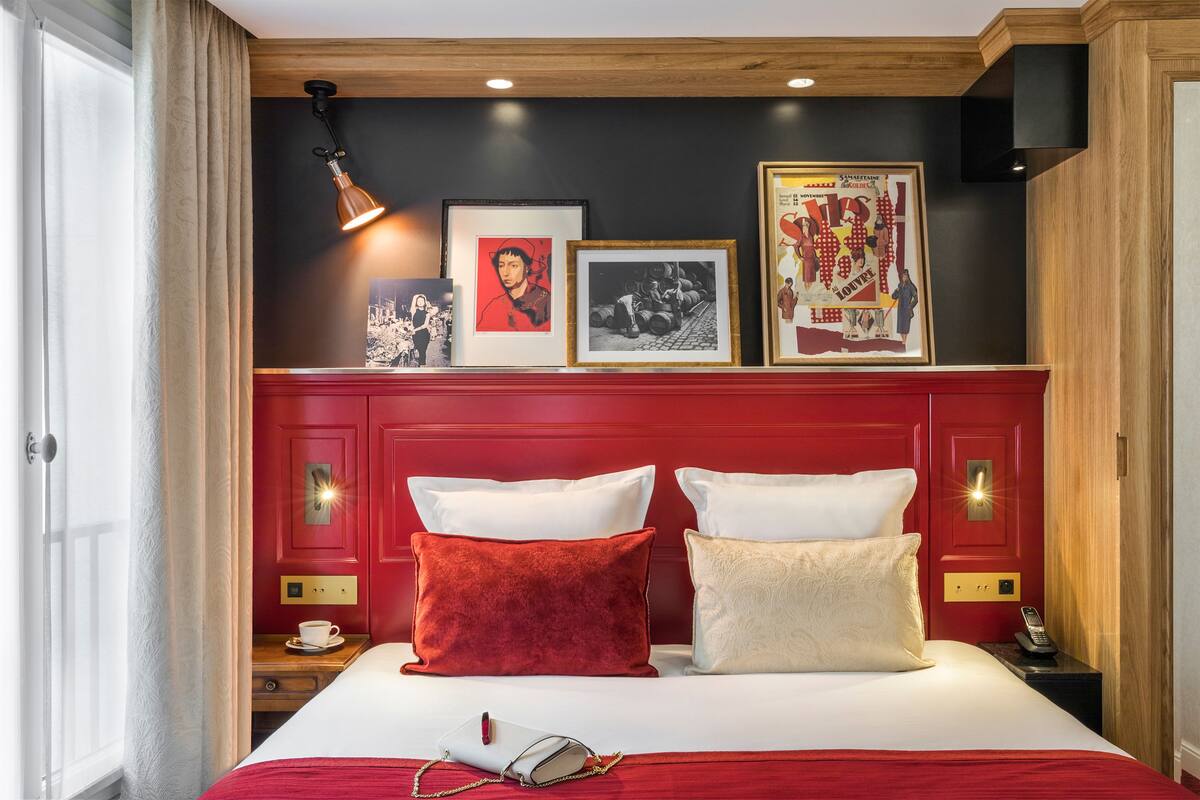 Une chambre d’hôtel avec une tête de lit rouge, une table de chevet sur laquelle se trouvent une tasse et un téléphone, et des œuvres d’art accrochées au mur.