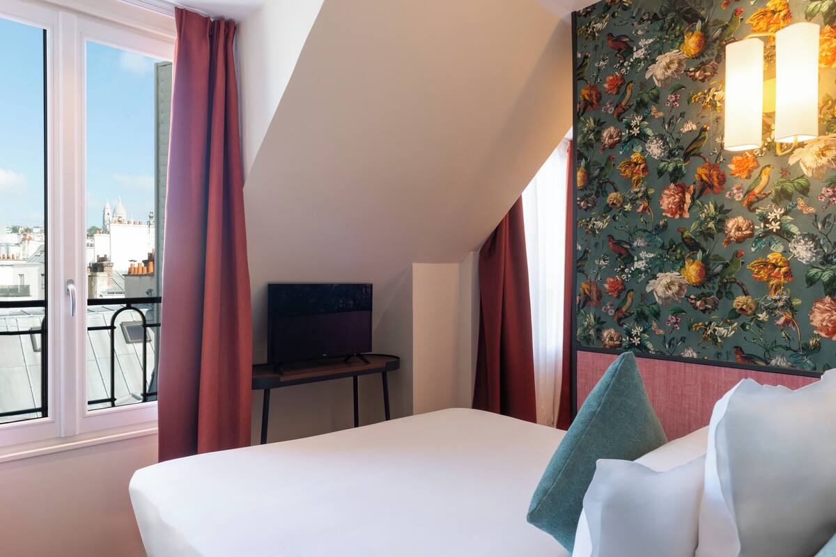 A hotel room with a large bed, a TV on a small table, and a balcony with a view of buildings.