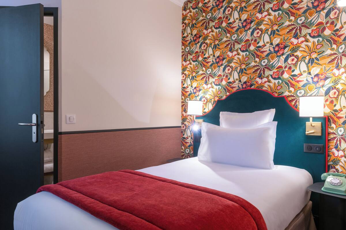 A hotel room with a bed, a red blanket, a floral wallpaper, and a telephone on the nightstand.