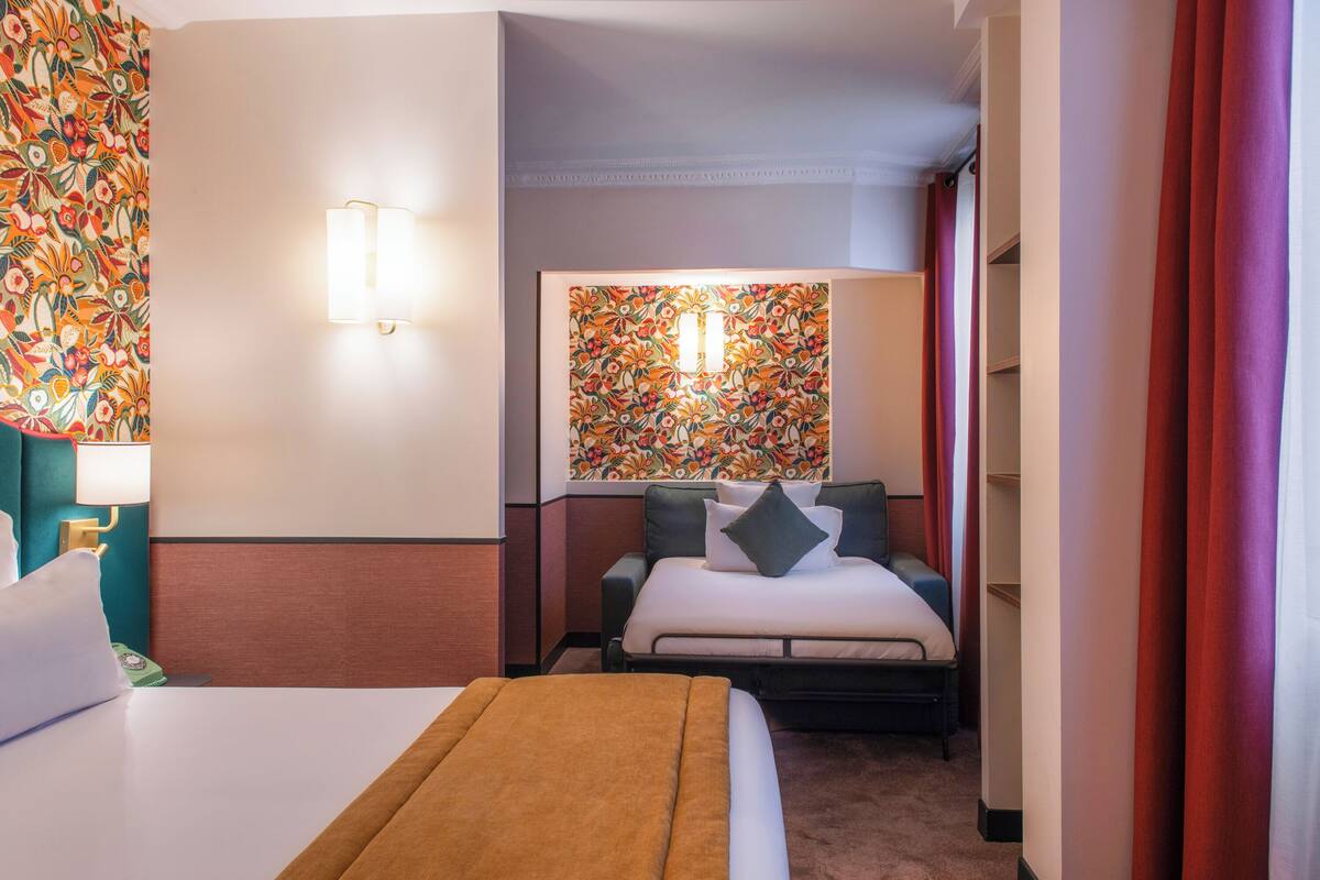 A hotel room with a bed, a bedside table, a lamp, a floral-patterned wall, and a window with curtains.