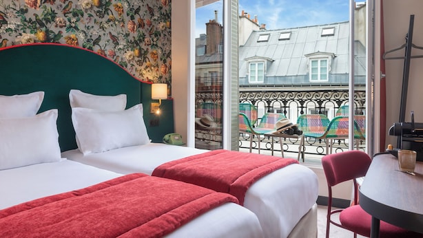 A hotel room with two beds, a balcony with a view of buildings, and a table with chairs. - Hôtel Brittany Opéra (Paris)