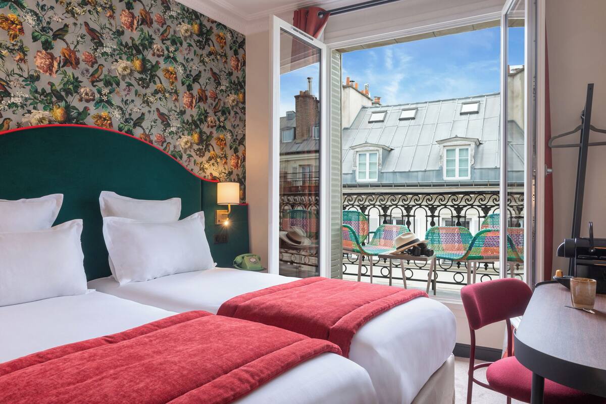 A hotel room with two beds, a balcony with a view of buildings, and a table with chairs.