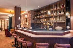 Uma área de bar com balcão de madeira, banquetas e prateleiras cheias de garrafas e copos.