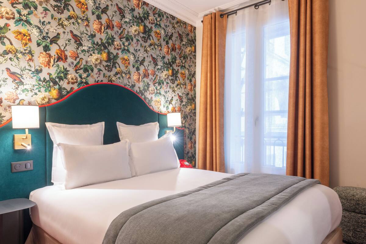 A hotel room with a floral wallpaper, a teal headboard, white bedding, and a view of the outdoors through a window with curtains.