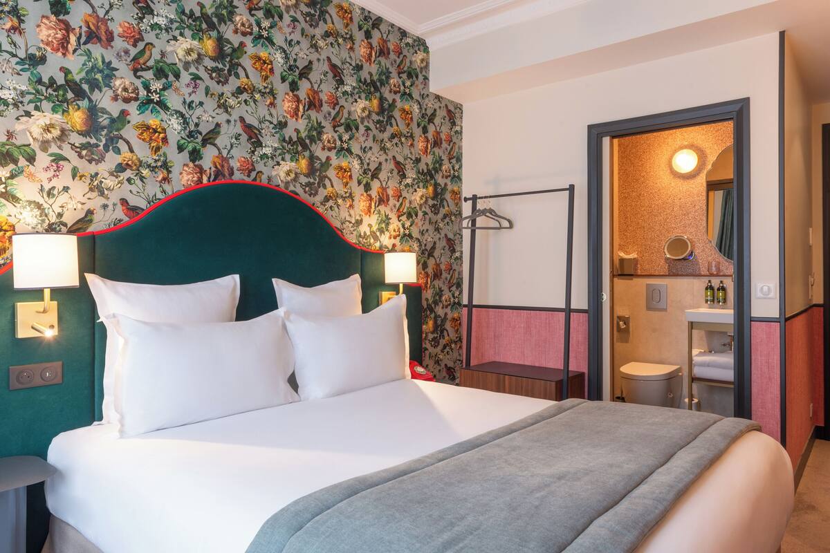 A hotel room with a large bed, floral wallpaper, a green headboard, bedside lamps, a small table, and a bathroom visible through an open door.