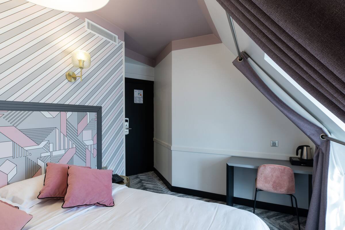 Habitación de hotel con cama, escritorio, silla, pared con patrones geométricos y ventana con cortinas.
