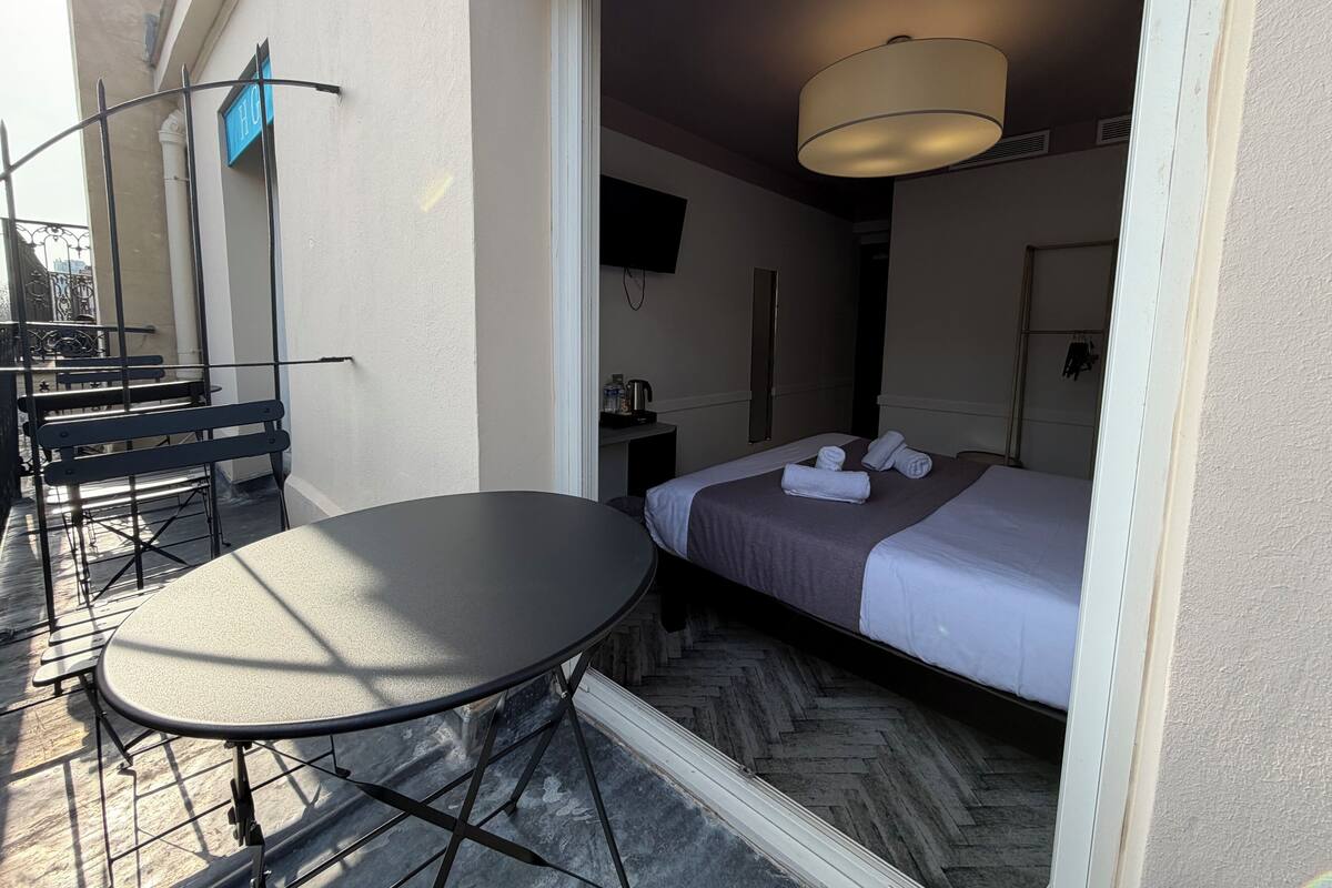 Habitación de hotel con cama, una mesa, una silla, televisión en la pared y balcón con mesa y sillas.