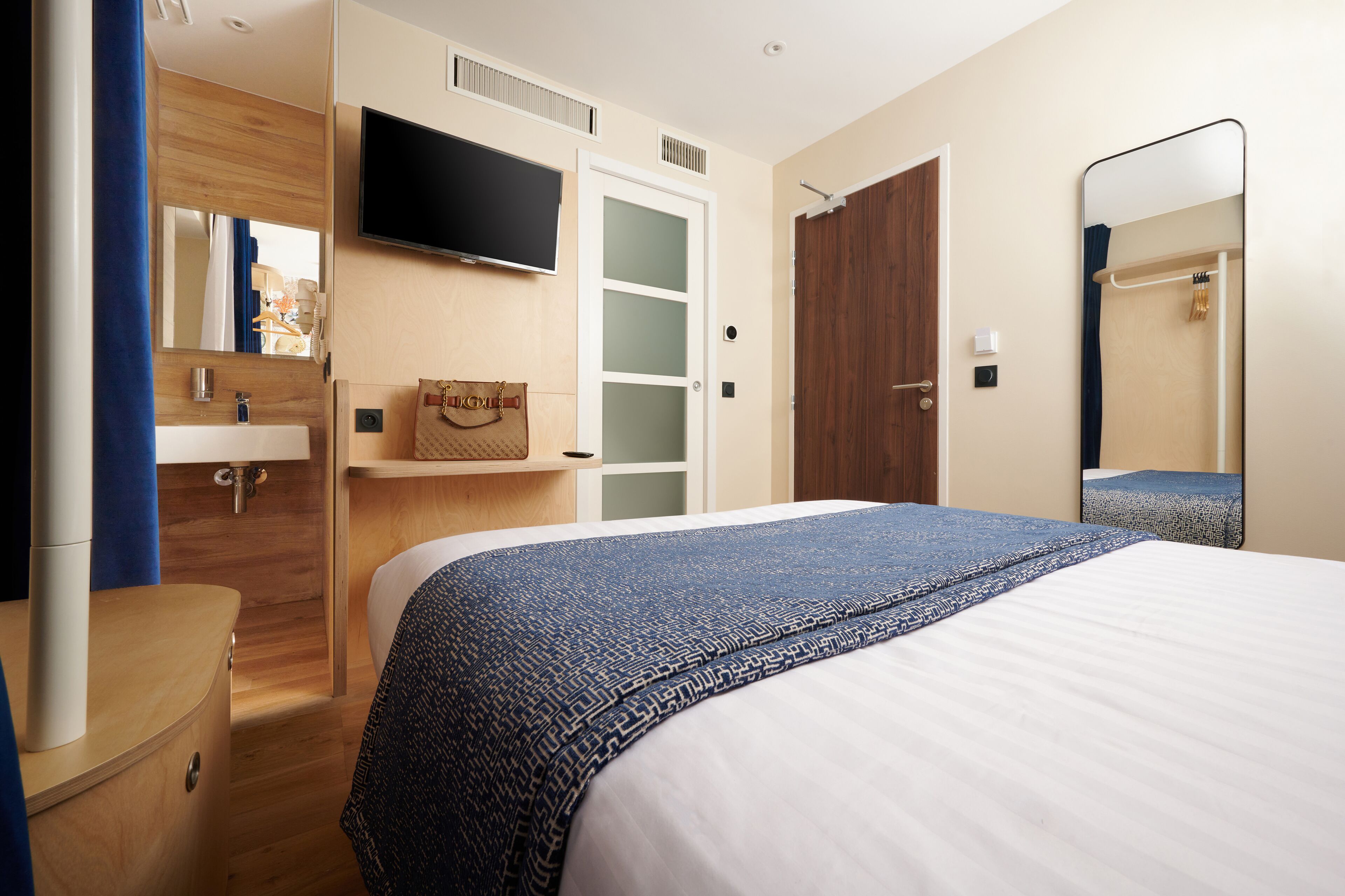 Une chambre d’hôtel avec un lit, un téléviseur fixé au mur, une commode en bois, une salle de bain avec un miroir et une porte.