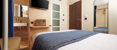Une chambre d’hôtel avec un lit, un téléviseur fixé au mur, une commode en bois, une salle de bain avec un miroir et une porte.