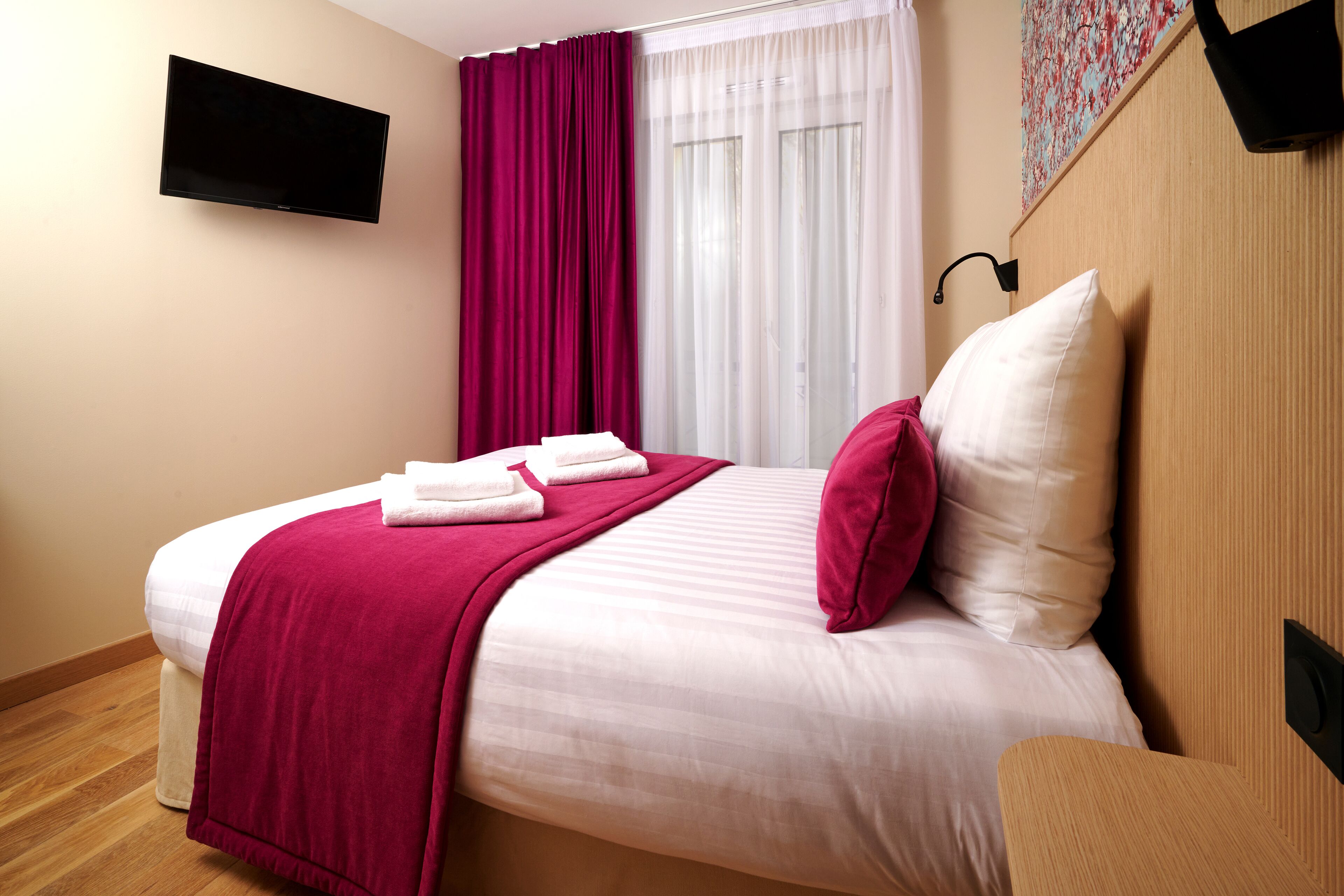Una cama bien hecha con una colcha blanca y rosa, una manta roja y dos toallas blancas. Un televisor de pantalla plana está montado en la pared sobre la cama.