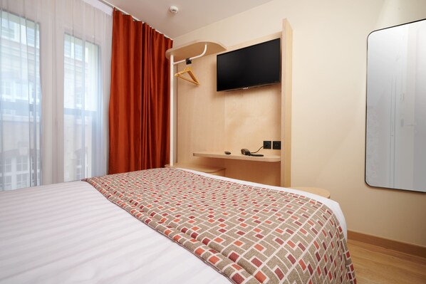Une chambre d’hôtel avec un lit, un téléviseur fixé au mur, un bureau en bois et une fenêtre avec des rideaux.