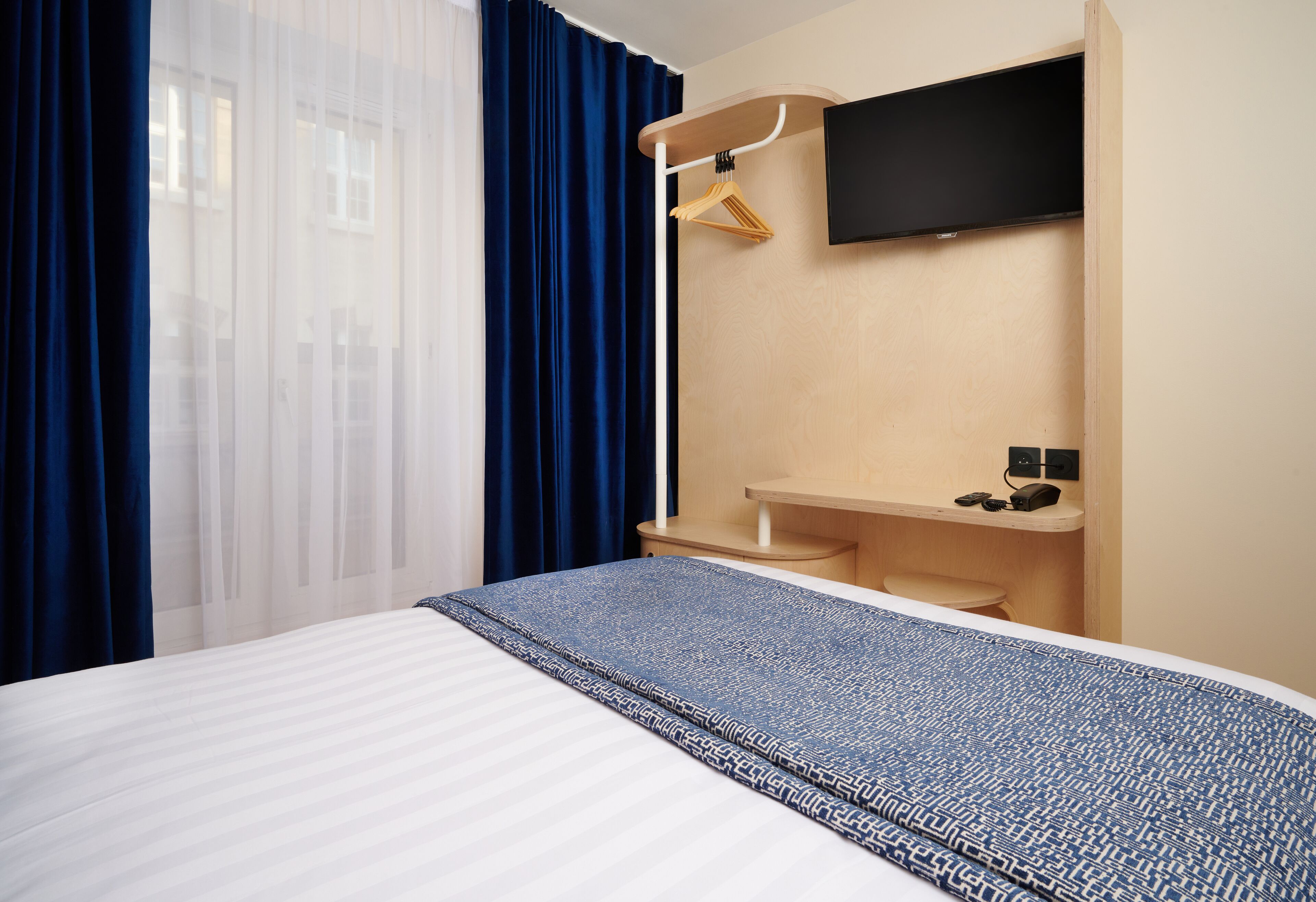 A hotel room with a bed, a desk, a chair, a TV mounted on the wall, and a window with curtains.