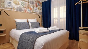A hotel room with a large bed, a wooden headboard, a patterned wall, and a window with blue curtains. - VLH Montparnasse Eiffel (Paris)