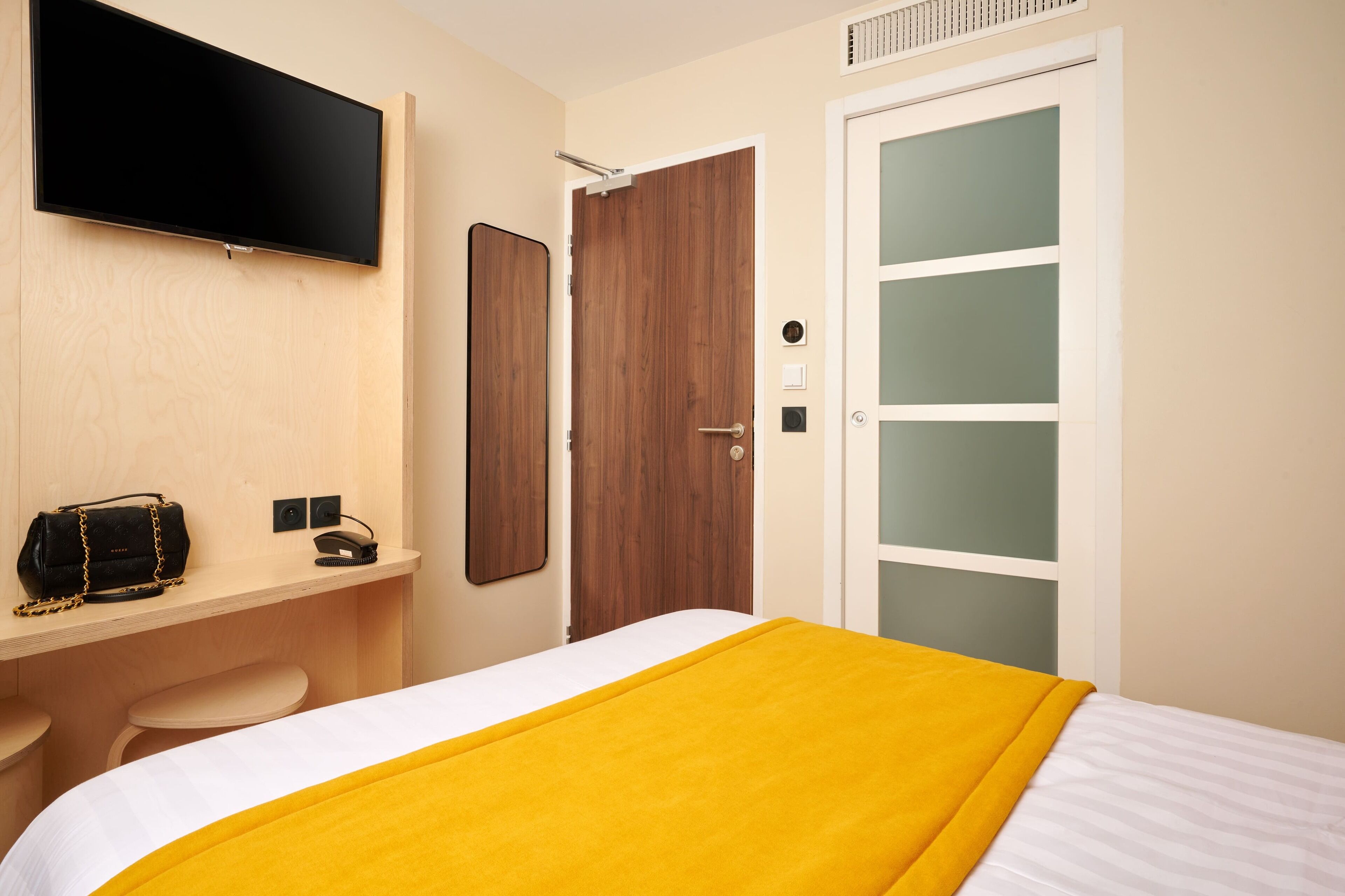 Quarto de hotel com cama, televisão fixada na parede, escrivaninha de madeira com uma bolsa e porta com painéis de vidro fosco.