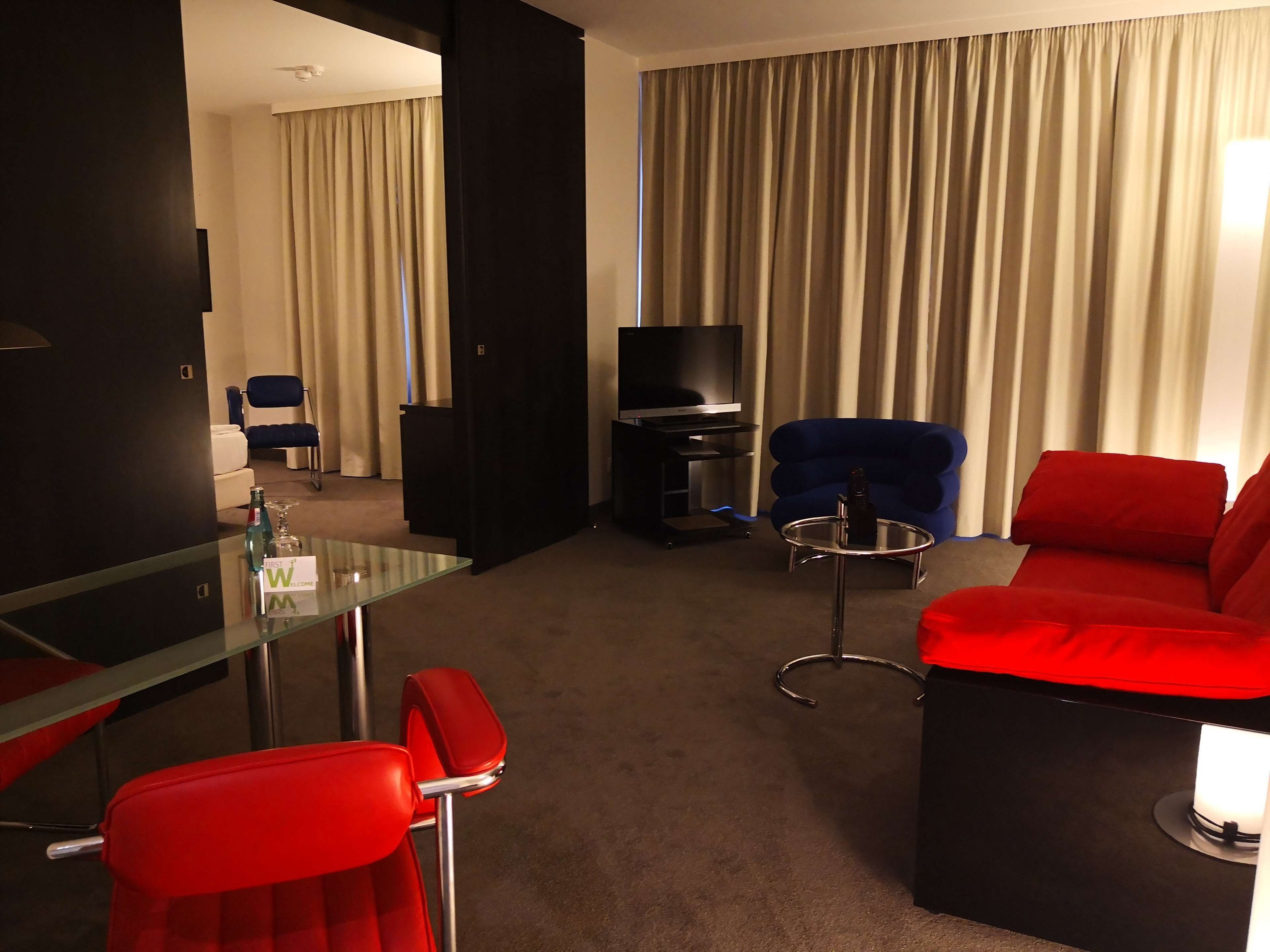 Habitación de hotel con una mesa de vidrio, sillas rojas, un sofá negro, un televisor y un ventanal grande con cortinas.