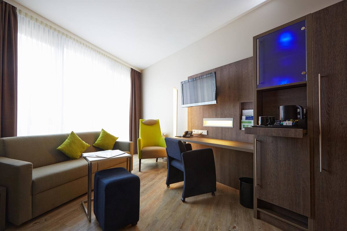 Una habitación de hotel moderna con sofá, sillones, una mesita, un escritorio, televisor y cafetera.