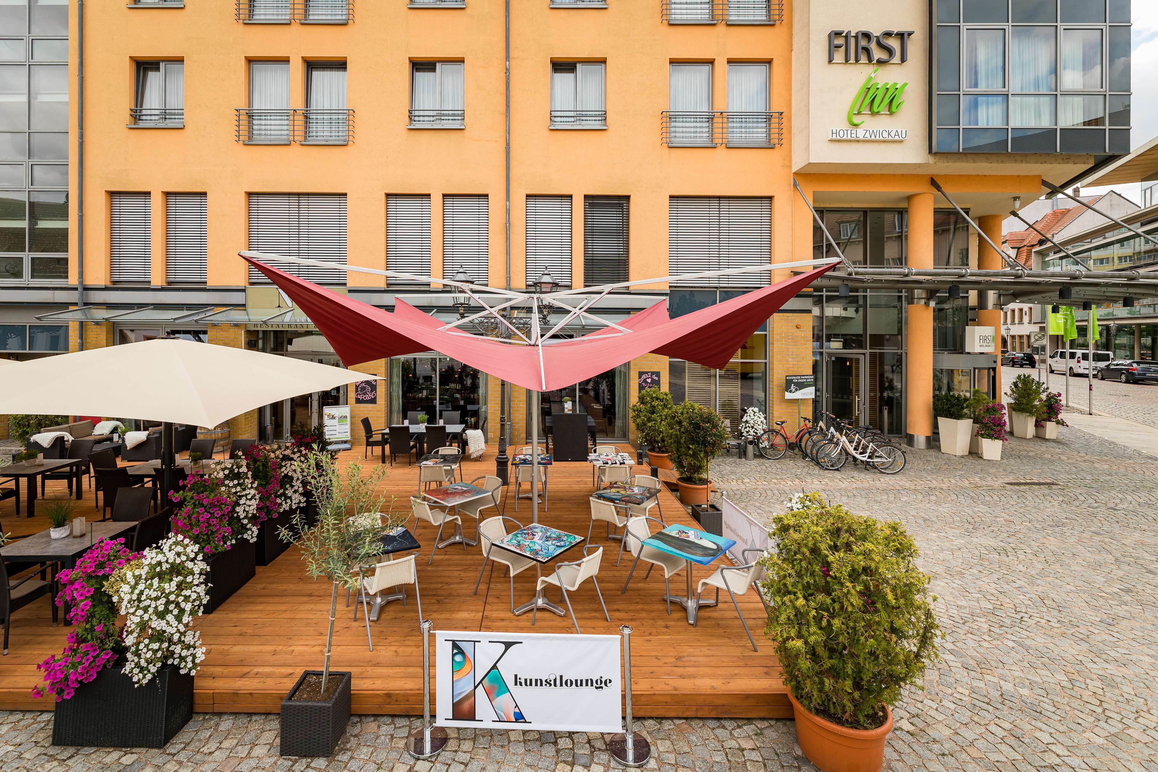 An outdoor seating area with tables and chairs, potted plants, and a large umbrella structure.