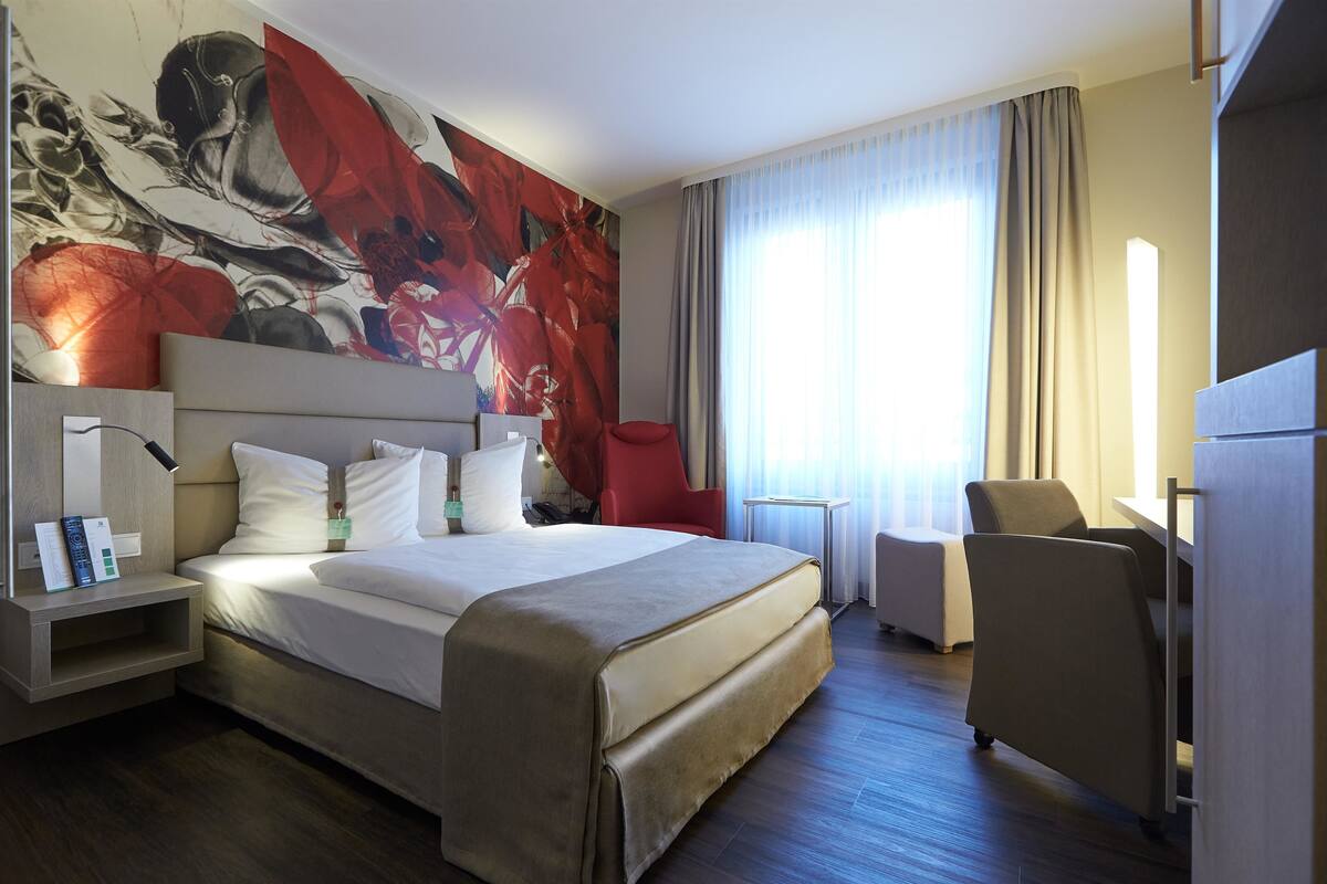 Una habitación de hotel moderna con una cama grande, una silla roja, un escritorio y una obra de arte en la pared.