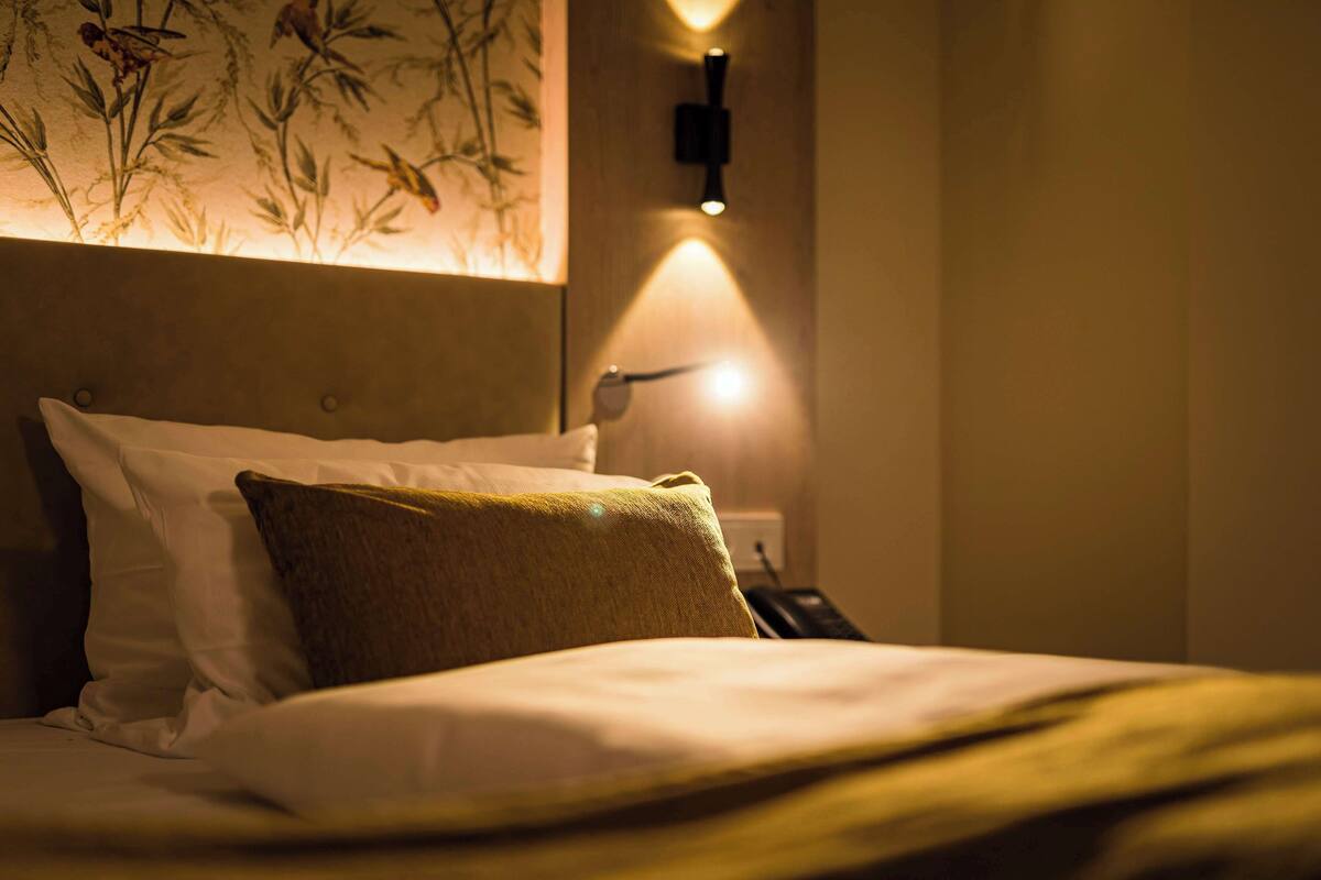 Una cama bien hecha con almohadas blancas y marrones, un aplique de pared y un estampado de papel tapiz floral.