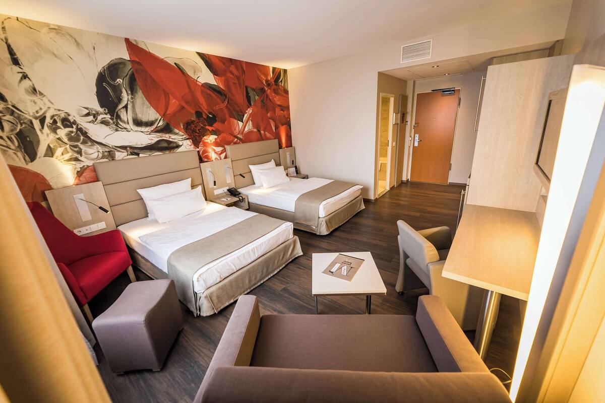 Una habitación de hotel con dos camas, una silla roja, un sofá marrón, una mesita pequeña y un cuadro abstracto grande en la pared.