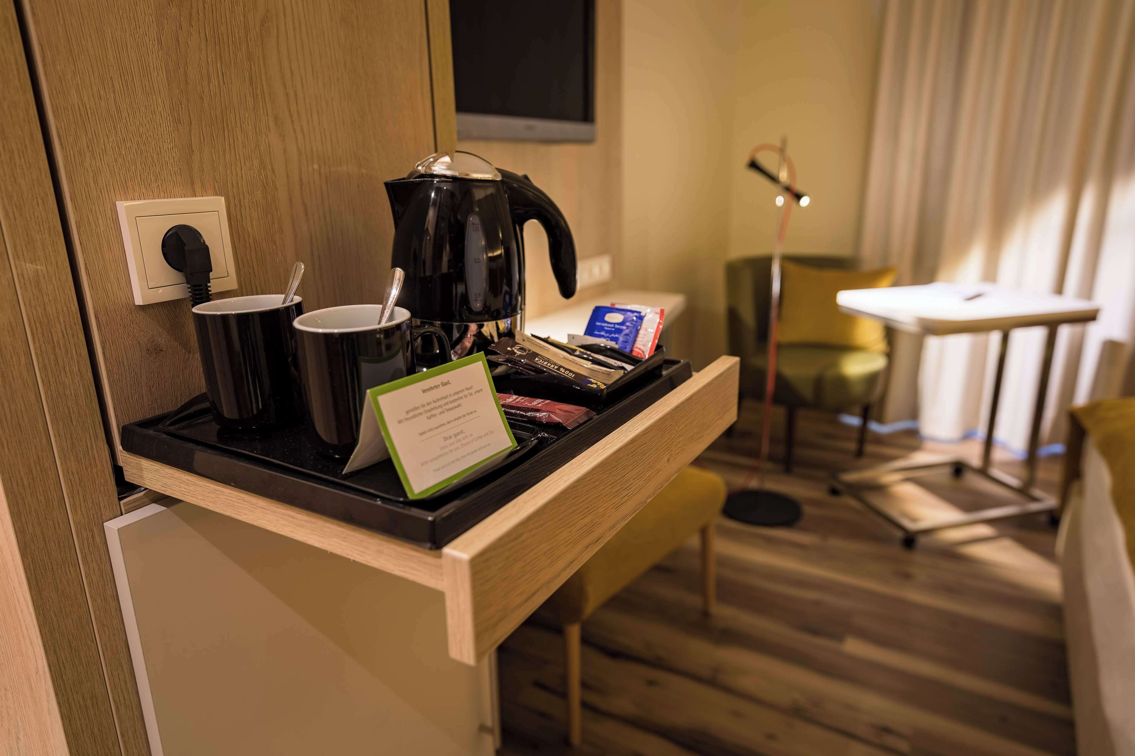 Habitación de hotel con estación de café, una mesita, una silla y un televisor montado en la pared.