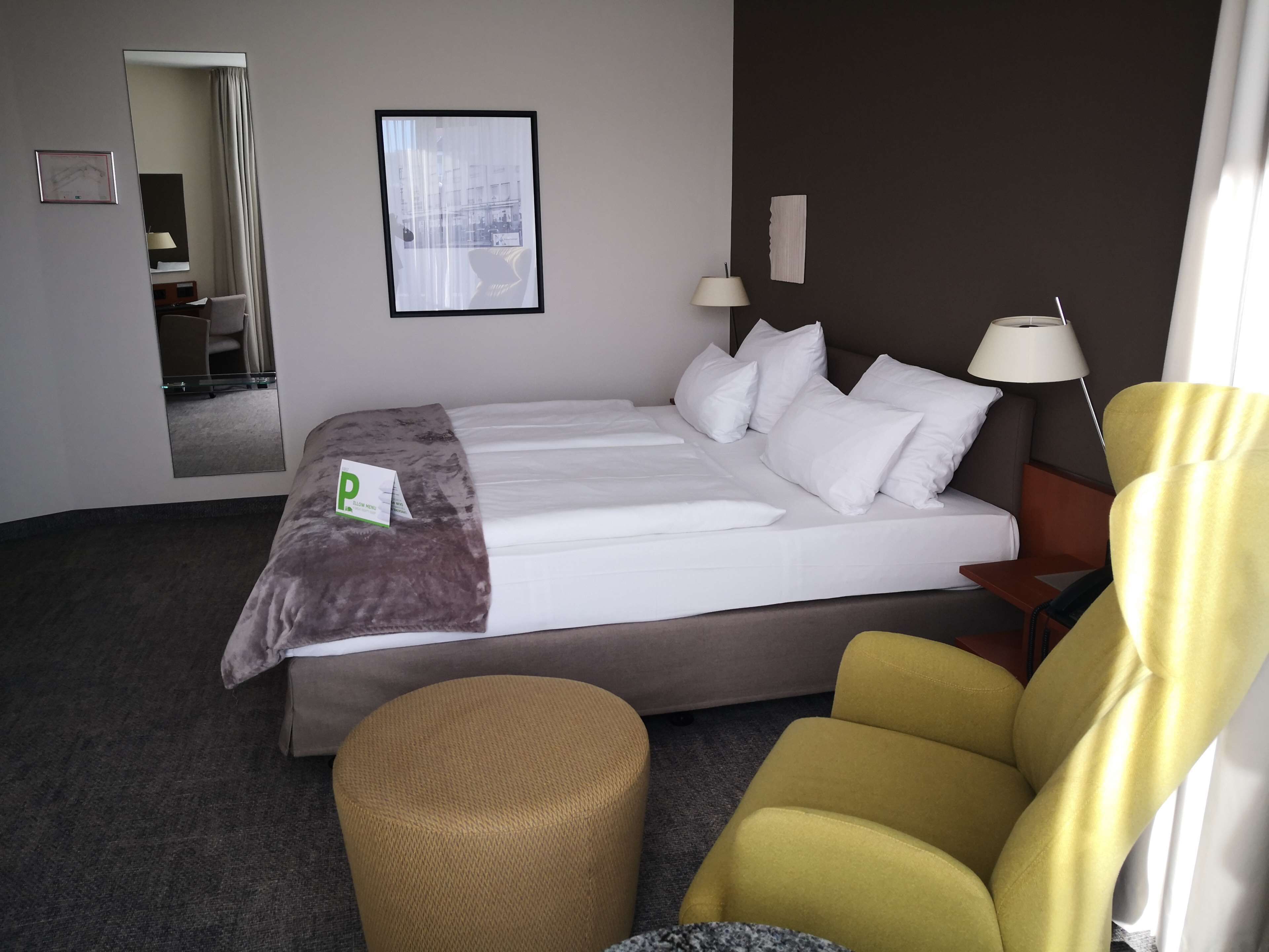 Een hotelkamer met een groot bed, een klein rond poefje, een gele fauteuil, een spiegel en een lijst met een afbeelding aan de muur.