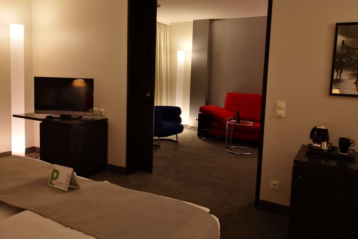 Una habitación de hotel con cama, televisor, escritorio, una silla roja y una pequeña mesa con cafetera.