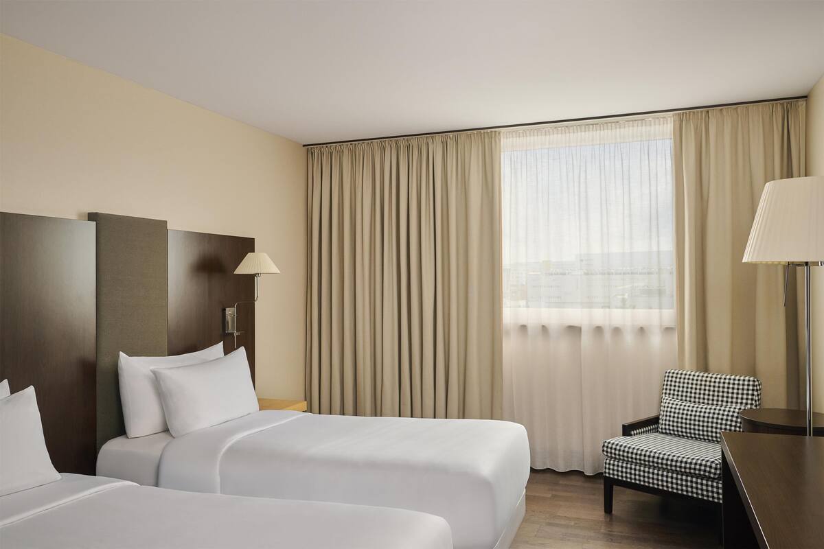 A hotel room with two beds, a checkered chair, a desk, and a large window with curtains.