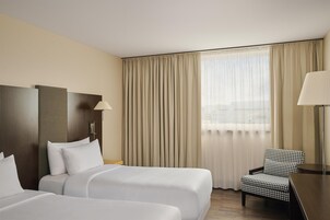 Quarto de hotel com duas camas, uma cadeira xadrez, uma mesa de trabalho e uma janela grande com cortinas.