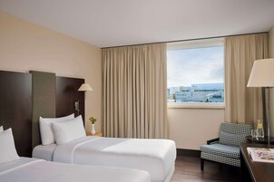 Habitación de hotel con dos camas, un sillón a cuadros, un escritorio con lámpara y vistas a la ciudad a través de amplias ventanas.