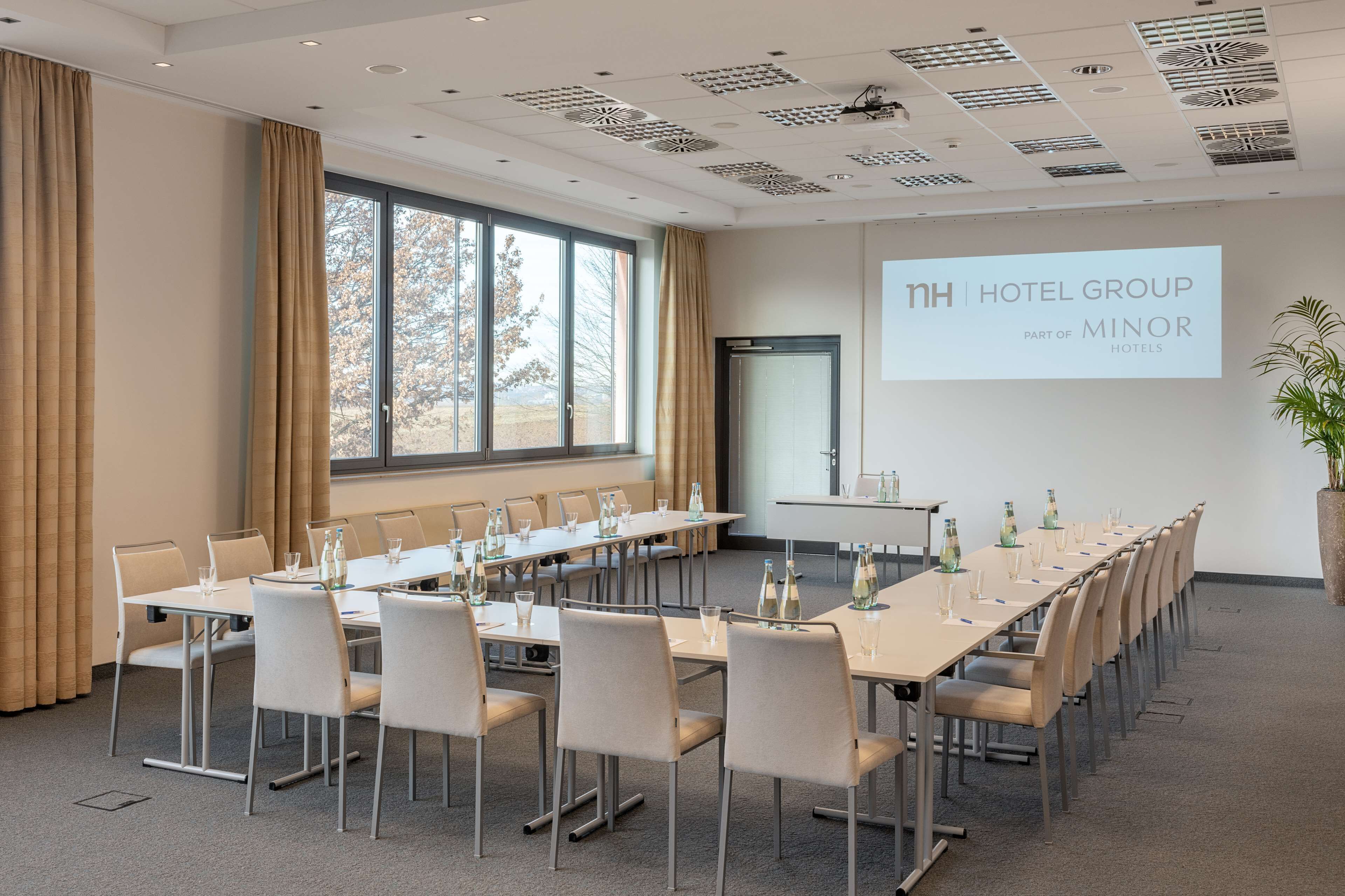 Una sala de conferencias con una mesa larga, sillas y una pantalla de proyector que muestra "nh HOTEL GROUP PARTE DE MINOR HOTELS".