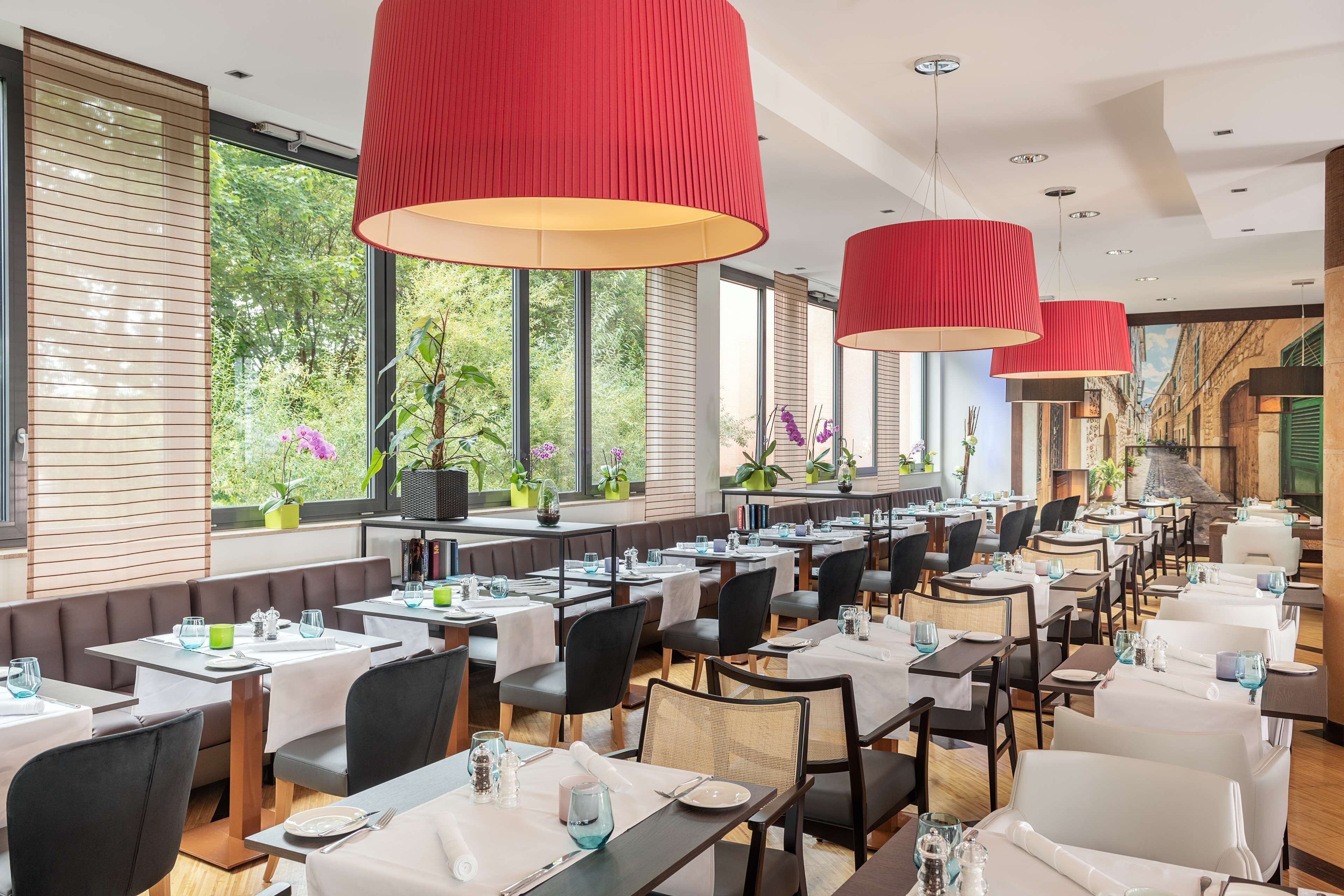 Un restaurante con varias mesas y sillas, grandes lámparas colgantes rojas y amplios ventanales con vista a zonas verdes.