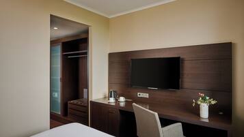 A hotel room with a bed, a desk with a chair, a TV mounted on the wall, a closet, and a small potted plant.