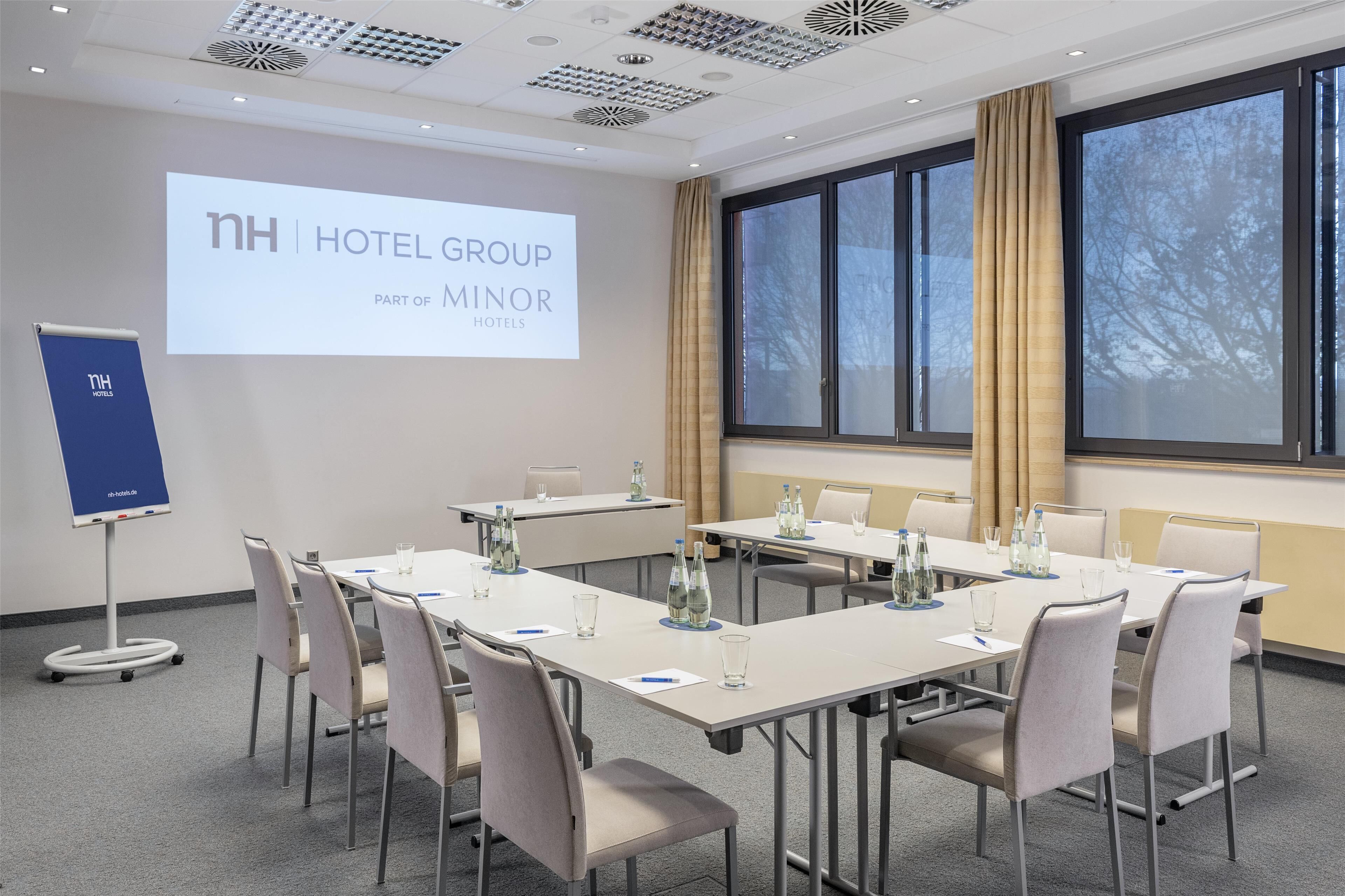 Un salón de conferencias de hotel con una pantalla grande que muestra “nh hotel group parte de minor hotels”, un proyector y un atril.
