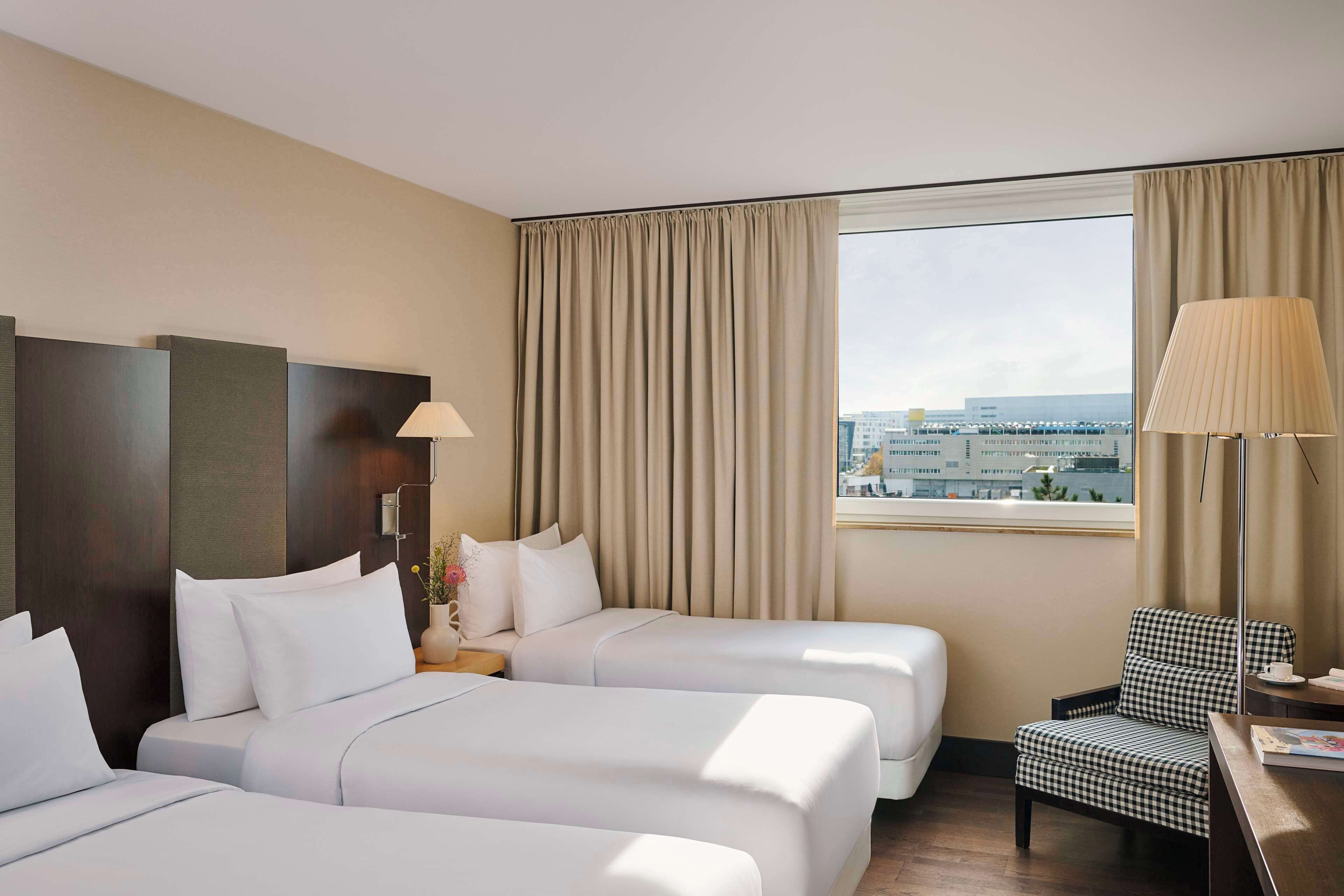 Une chambre d’hôtel avec deux lits, un bureau avec une lampe, une chaise et une grande fenêtre avec des rideaux.