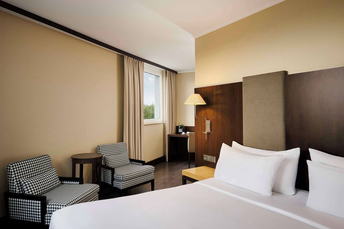 A hotel room with a bed, two checkered armchairs, a desk, a lamp, and a window with curtains.