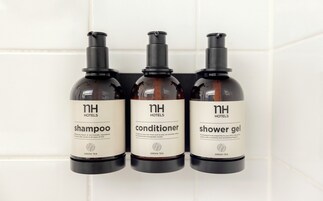 Tres botellas de artículos de tocador de hotel con la etiqueta 'NH Hotels' sobre una pared de azulejos.