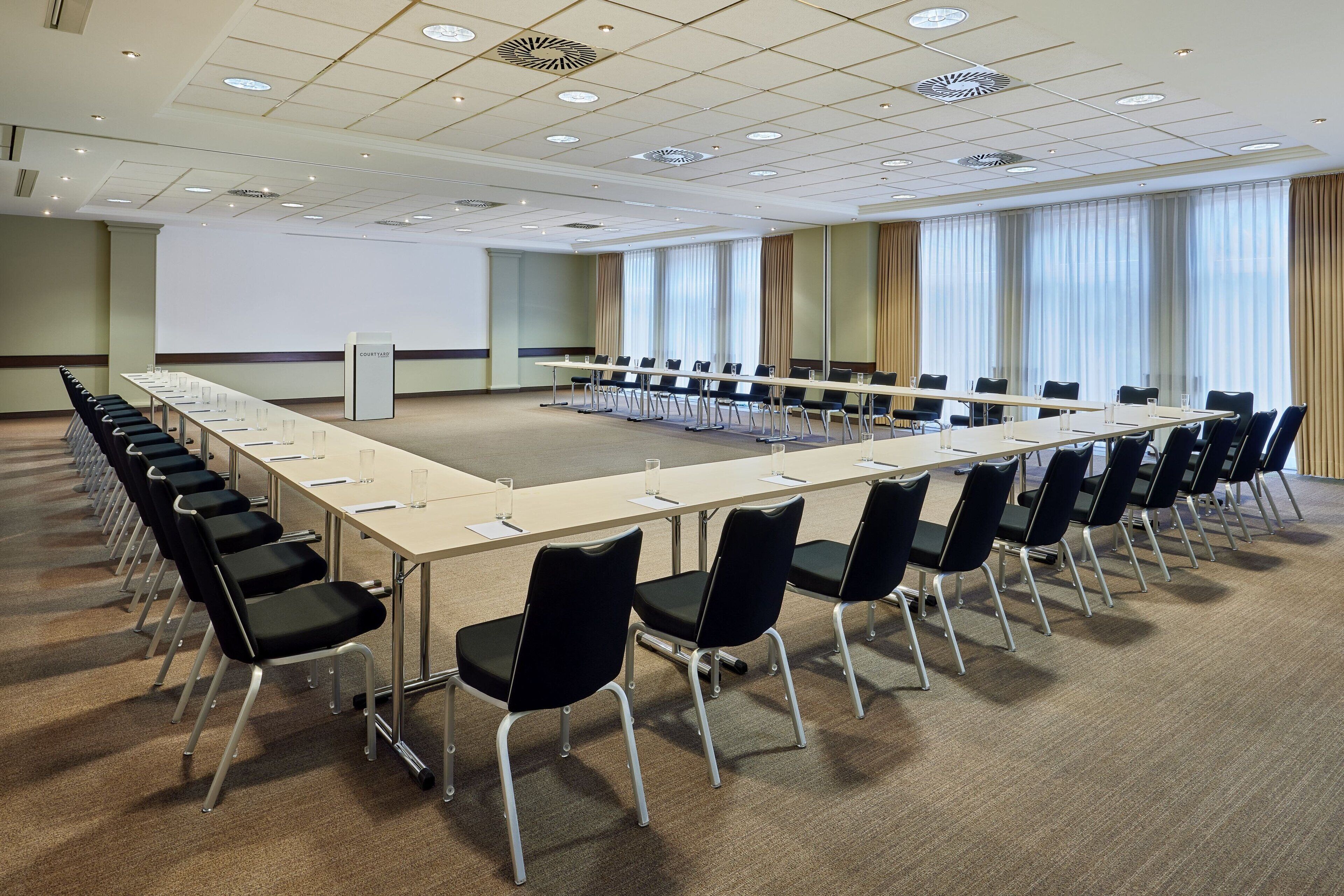 Une salle de conférence avec une longue table, des chaises noires, un écran de projection et de grandes fenêtres équipées de rideaux.