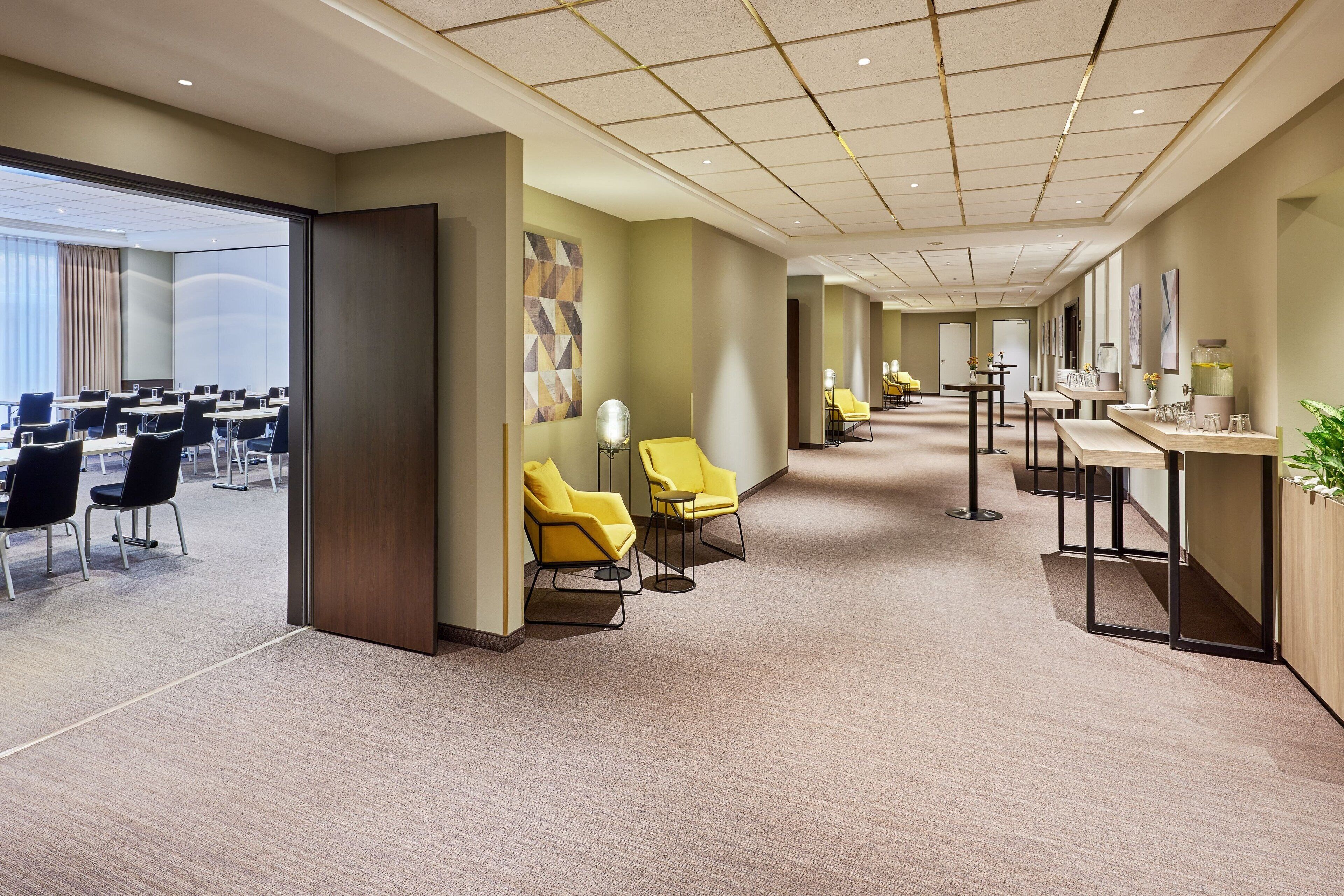 A hotel lobby with a long corridor, yellow chairs, a reception desk, and a large window with curtains.