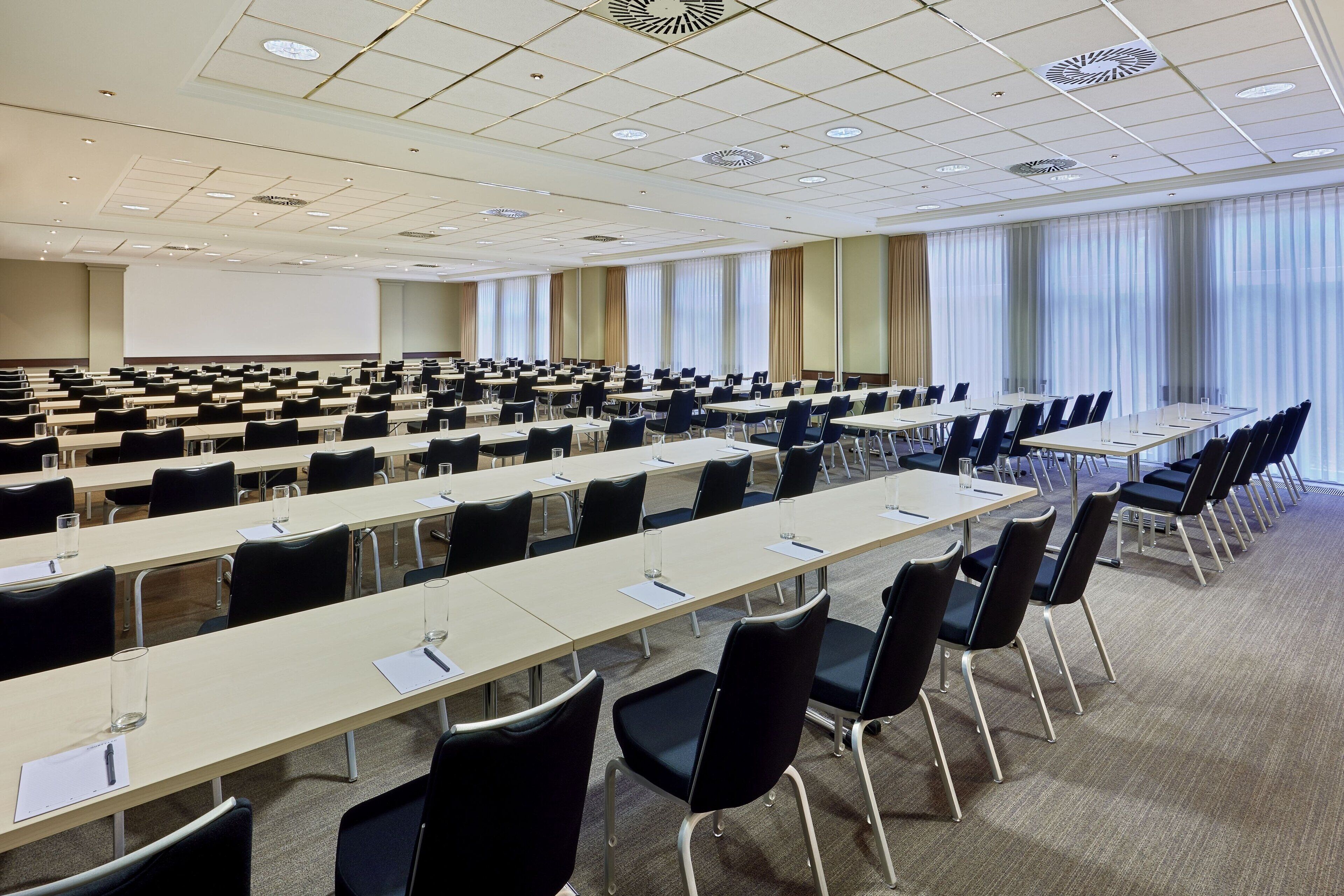 A large conference room with rows of tables and chairs, a projector screen, and large windows with curtains.