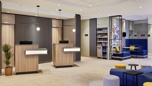 A modern reception area with two wooden reception desks, a blue seating area with yellow cushions, and a vending machine. - Courtyard by Marriott Schwerin (Schwerin)